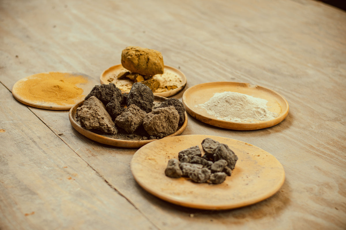 Raw clay minerals displayed on wooden dishes — bentonite chunks, kaolin powder, and earth minerals used in the NINÜ Full Spectrum Detox Clay Bath Soak