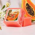 Papaya scented soap bar in a clear geometric box with a printed label, shown next to a loose soap bar, with a sliced papaya and white flowers in the background.