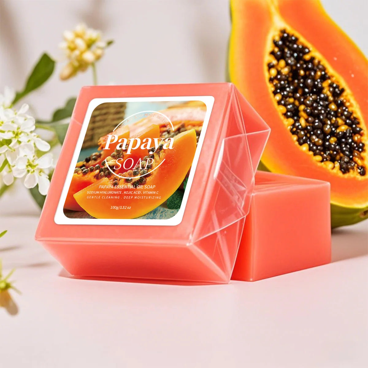 Papaya scented soap bar in a clear geometric box with a printed label, shown next to a loose soap bar, with a sliced papaya and white flowers in the background.